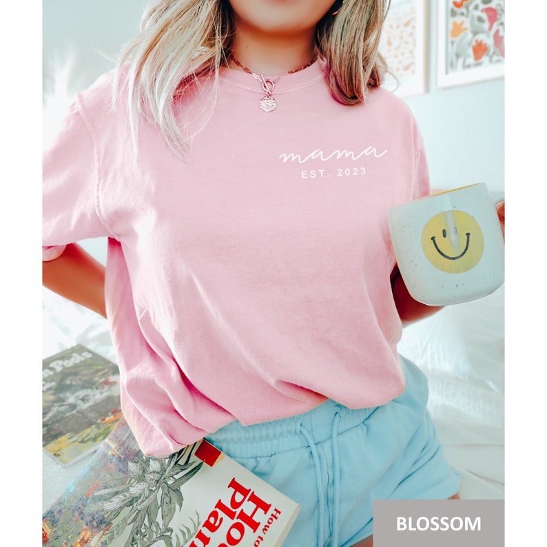 Pu&ograve; includere: Una maglietta rosa con il testo "mama est. 2023" in bianco. La persona che indossa la maglietta tiene una tazza bianca con una faccia sorridente gialla.