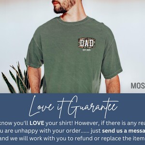 May include: Moss green t-shirt with a camouflage-style "DAD" graphic and "EST. 2025" text. The shirt is a solid color and has a crew neck. The word "MOSS" is displayed in the lower right corner.