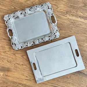 Peut inclure: Deux plateaux de service argentés sur une surface en bois. L'un des plateaux est de forme rectangulaire avec des poignées et un motif de vigne sur les bords. L'autre plateau est rectangulaire avec des poignées et un design simple.