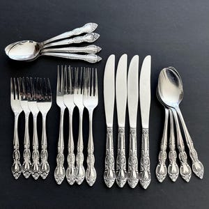 May include: A set of silver-coloured cutlery, including spoons, forks, and knives, arranged on a black surface. The silverware features ornate floral detailing on the handles. The set includes multiple forks, knives, and spoons.