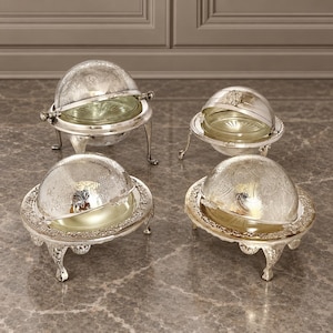 May include: Four silver-plated serving dishes with rotating dome lids and glass inserts. Each piece features ornate engraved patterns on the metal covers, decorative scalloped edges, and stands on three curved legs with intricate detailing.
