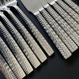 May include: Close-up of a set of stainless steel cutlery. The forks and knives have a textured, woven pattern on the handles. The cutlery is arranged diagonally against a dark background, highlighting the reflective quality of the metal.