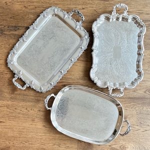 Peut inclure: Trois plateaux de service en métal argenté avec des poignées ornées et des bordures décoratives en relief. Les plateaux rectangulaires et ovales présentent des motifs floraux et des volutes complexes gravés sur leurs surfaces réfléchissantes.