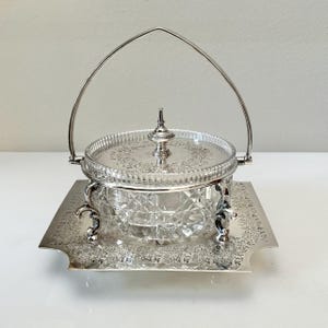 May include: A silver-plated serving dish with a clear, diamond-cut glass bowl and lid. The dish has a decorative handle and sits on a matching tray with ornate floral detailing. The design is elegant and vintage.