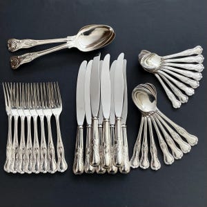 May include: A collection of antique silverware, including forks, knives, and spoons, displayed on a dark background. The silverware features decorative floral patterns on the handles and a polished, reflective finish. The spoons and knives are arranged in a fan-like pattern.