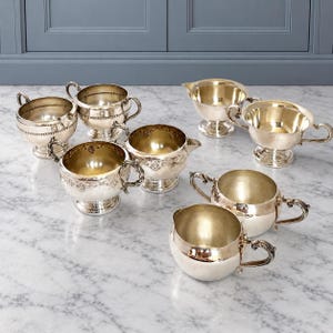 May include: A collection of seven silver-toned metal bowls and creamers, each featuring a pedestal base and two side handles. Some pieces have smooth surfaces, while others display intricate embossed floral and beaded decorative details.