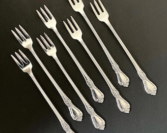 May include: A collection of eight silver-colored dessert forks arranged diagonally on a dark surface. Each fork has three tines and a decorative handle with floral detailing. The forks are made of a shiny, reflective metal.