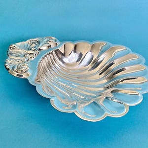 May include: A polished silver-toned metal dish shaped like a seashell. The dish features a scalloped edge and a decorative, embossed hinge detail at the top, with a smooth, reflective surface that catches light across its curved, fluted ridges.