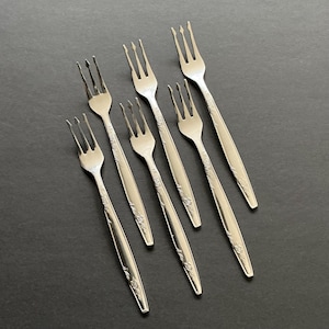 May include: Six silver forks with a floral design on the handles are arranged on a dark surface. The forks have three tines and are likely for dessert or appetizers. The silverware is likely vintage.