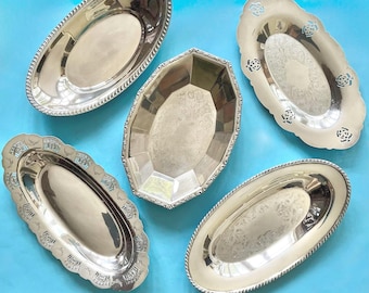 May include: Five silver serving trays of various shapes and designs are arranged on a light blue surface. The trays include oval, rectangular, and faceted shapes, with decorative edges and embossed patterns. The trays are reflective and appear to be made of metal.