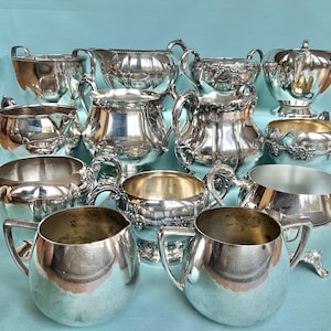 May include: An assortment of antique silver serving pieces, including pitchers, sugar bowls, and cream jugs. The items feature intricate detailing and are arranged on a light blue surface. The silver reflects light, creating a bright and elegant display.