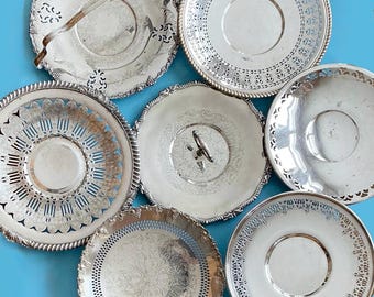 May include: A collection of six circular silver-toned metal serving trays and saucers. Each piece features intricate openwork lattice borders, embossed floral patterns, and smooth central surfaces. One tray includes a central handle.