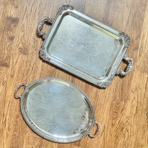 May include: A rectangular silver-coloured serving tray with ornate handles and an embossed floral centre, paired with an oval silver-coloured serving tray featuring decorative handles and a repeating circular pattern engraved on its surface.