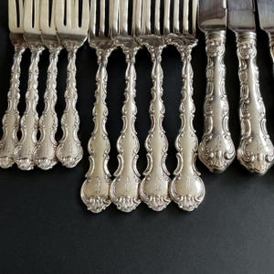 May include: Close-up of antique silverware, including forks and knives, arranged on a dark surface. The silverware has ornate, detailed handles with a floral design. The forks and knives are made of a shiny, silver-coloured metal.