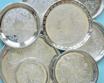 May include: A collection of round silver-toned metal trays featuring intricate embossed floral and scrollwork patterns along the borders and centers. The trays have raised, decorative edges and polished surfaces with reflective finishes.