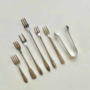 May include: A collection of six silver-toned metal utensils including five small forks of varying lengths and designs, some with floral embossed handles, and one pair of U-shaped serving tongs with curved ends, all arranged in a diagonal row.