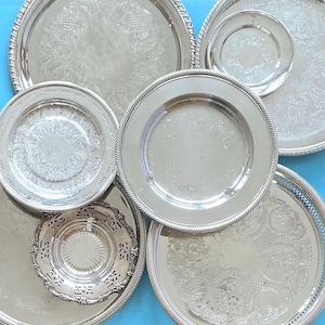 May include: A collection of six round silver-toned metal serving trays and plates of varying sizes. The items feature diverse surface textures, including intricate embossed floral patterns, smooth polished centres, and decorative rope-style edges.