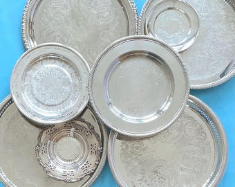 May include: A collection of six round silver-toned metal serving trays and plates of varying sizes. The items feature diverse surface textures, including intricate embossed floral patterns, smooth polished centers, and decorative rope-style edges.