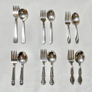 May include: Set of six silver-plated baby forks and spoons. The forks have three tines and the spoons have a rounded bowl. The handles are ornate and feature a floral design.