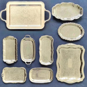 Peut inclure: Un ensemble de neuf plateaux de service en métal argenté de formes et tailles variées, incluant des modèles rectangulaires, ovales et festonnés. Chaque plateau présente des motifs floraux en relief et des surfaces polies avec des poignées.