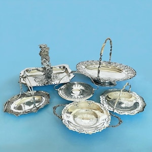May include: A selection of ornate silver serving dishes and trays. The collection includes trays with handles, decorative edges, and a tray with a grape vine handle. The pieces are displayed on a light blue background.