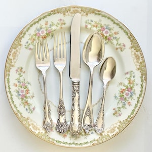 Peut inclure: Un ensemble de cinq couverts en métal argenté comprenant deux fourchettes, un couteau, une cuillère à soupe et une petite cuillère, disposés verticalement sur une assiette en céramique à fond crème avec une bordure de fleurs et de vignes.