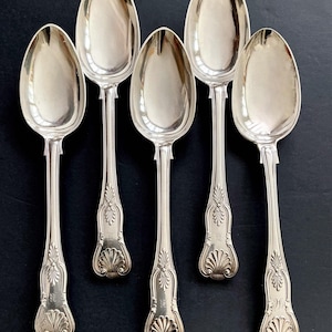 May include: Six silver spoons with ornate handles and a reflective surface. The spoons have a shell design at the base of the handle and a detailed floral pattern. The spoons are arranged on a dark surface.