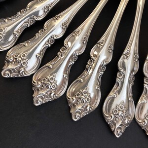 May include: Close-up of a collection of ornate silver-coloured forks. The handles feature a floral design with intricate details. The forks are arranged on a dark surface, showcasing their elegant, vintage style.