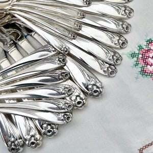 May include: A close-up of a collection of silver cutlery, including spoons and forks, with floral patterns on the handles. The silverware is arranged in a pile, with some pieces overlapping. A white cloth with floral embroidery is in the background.