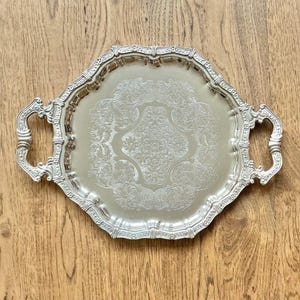 May include: A silver-coloured metal serving tray with an octagonal shape and two ornate handles. The centre of the tray features an intricate embossed floral and scrollwork pattern, while the raised rim has a decorative scalloped edge design.