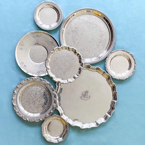 May include: A selection of antique silver serving trays and plates in various sizes and designs. The trays and plates feature decorative edges and embossed patterns, arranged on a light blue surface.