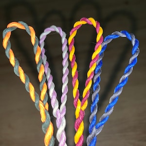 Może przedstawiać: Cztery skręcone sznurki w różnych kolorach: pomarańczowy i zielony, biały i fioletowy, żółty i bordowy oraz niebieski i szary.
