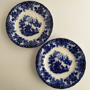 May include: Two blue and white porcelain plates with a decorative floral border and a central design of a pagoda and trees in a landscape setting.