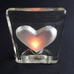 Può includere: Un blocco di vetro trasparente con un cuore argentato all'interno. Il cuore è illuminato da dietro, creando un bagliore caldo.
