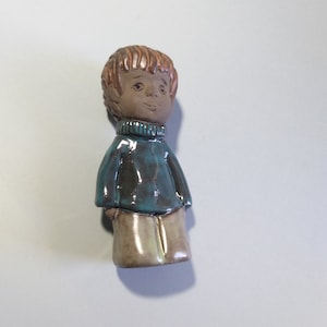 May include: A ceramic figurine of a boy with brown hair, wearing a teal sweater and beige pants.