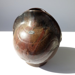May include: A brown ceramic vase with a unique, organic shape and a textured, mottled glaze. The vase has a small, round opening at the top and a wide, rounded base. It features a small, circular label with the letter "V" inside.