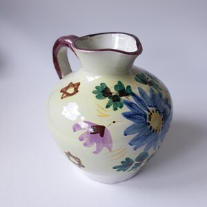 May include: A small, white ceramic jug with a purple, blue and green floral design. The jug has a brown handle and a Star of David design on the side.