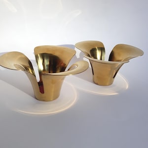 Op de afbeelding: Twee messing bloemvormige kandelaars. Elke houder heeft een uitlopend, bloembladachtig ontwerp met een cirkelvormige basis. De metallic gouden afwerking reflecteert licht, waardoor een warme uitstraling ontstaat. Deze decoratieve stukken zijn ideaal.