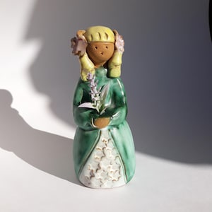May include: A ceramic figurine of a girl wearing a green dress with white flowers and a yellow crown. She is holding a lavender flower in her hands.