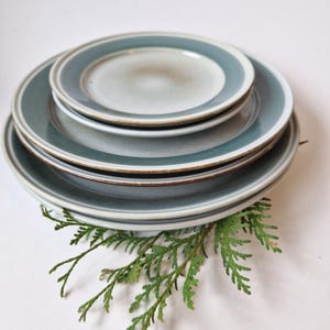Può includere: Una pila di piatti in ceramica di varie dimensioni. I piatti hanno un bordo blu-azzurro e un centro beige chiaro. Un rametto di fogliame verde si trova nella parte inferiore della pila. I piatti sono impilati su una superficie bianca.