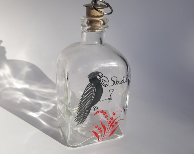Smålands Alster Kosta Sweden small bottle with a rectangular bottom. One side depicts a crow with a glass. Clear glass.