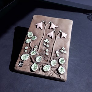 May include: A brown ceramic wall plaque with a raised design of pink flowers and green leaves. The plaque is signed "jic" in the bottom left corner.