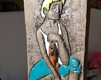May include: A ceramic wall sculpture of a person with blonde hair wearing a blue and white outfit. The sculpture is painted with brown, blue, white, yellow, and orange colors.