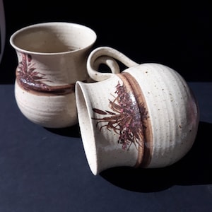 Può includere: Due tazze in ceramica con una glassa marrone macchiata e un motivo floreale marrone e rosa. Le tazze sono fatte a mano e hanno un aspetto rustico.