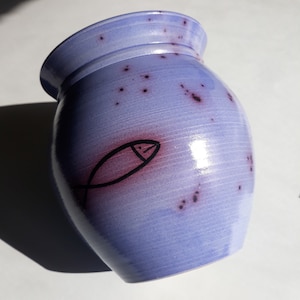 Peut inclure: Un vase en céramique bleu clair avec un motif de poisson noir peint à la main. Le vase a un glaçage violet moucheté et une forme arrondie.