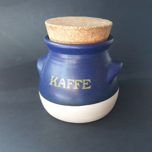Può includere: Un barattolo in ceramica blu e bianco con un coperchio di sughero. Il barattolo ha la scritta "Kaffee" in lettere dorate.