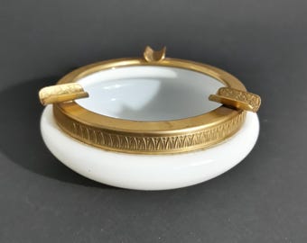 May include: A white ceramic ashtray with a gold-colored metal rim and accents. The ashtray has a circular design with a white interior and a decorative pattern around the base. It features three rests for cigarettes.