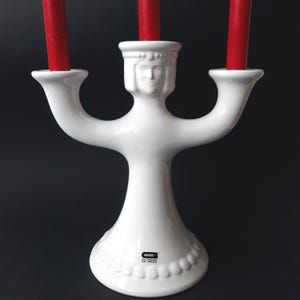 Könnte beinhalten: Ein weißer Kerzenhalter aus Keramik mit drei Armen, die jeweils eine rote Kerze halten. Der Halter hat ein stilisiertes Gesicht und einen dekorativen Sockel. Das Stück ist mit einem Logo mit der Aufschrift "GOLDBERGS ART SWEDEN" versehen.