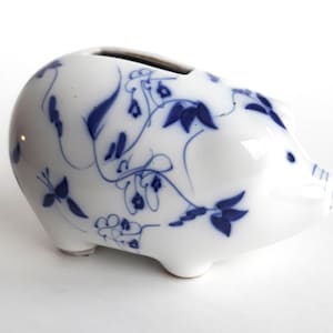 May include: A white ceramic piggy bank with a blue floral design. The piggy bank has a slot on the top for coins.