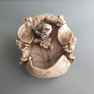 May include: A ceramic wall hanging featuring a bird and leaf design. The bird is light blue and white, and the leaves are brown. The wall hanging has a small, indented area in the centre.
